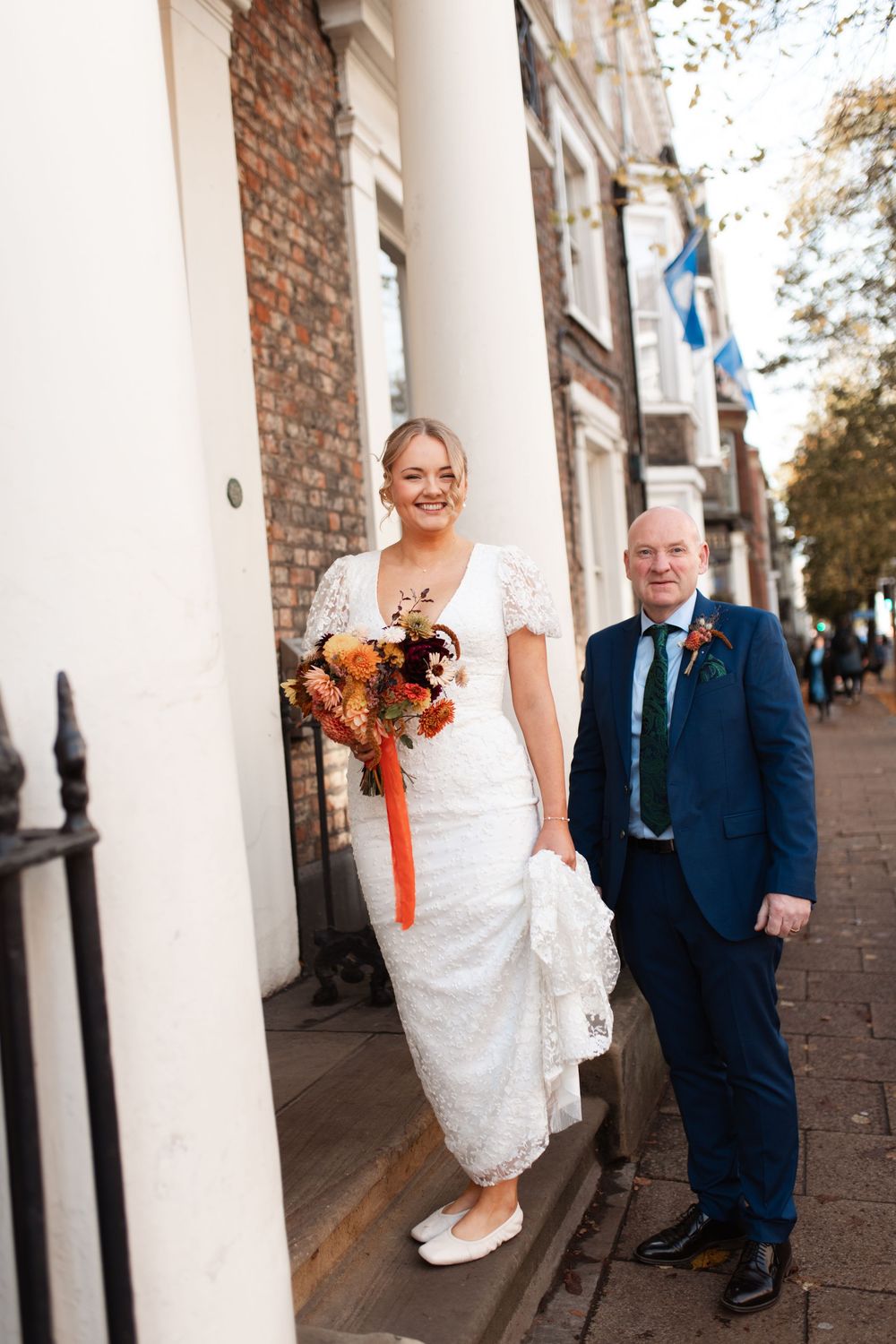 A couple poses between white columns against a brick wall during their wedding photoshoot.