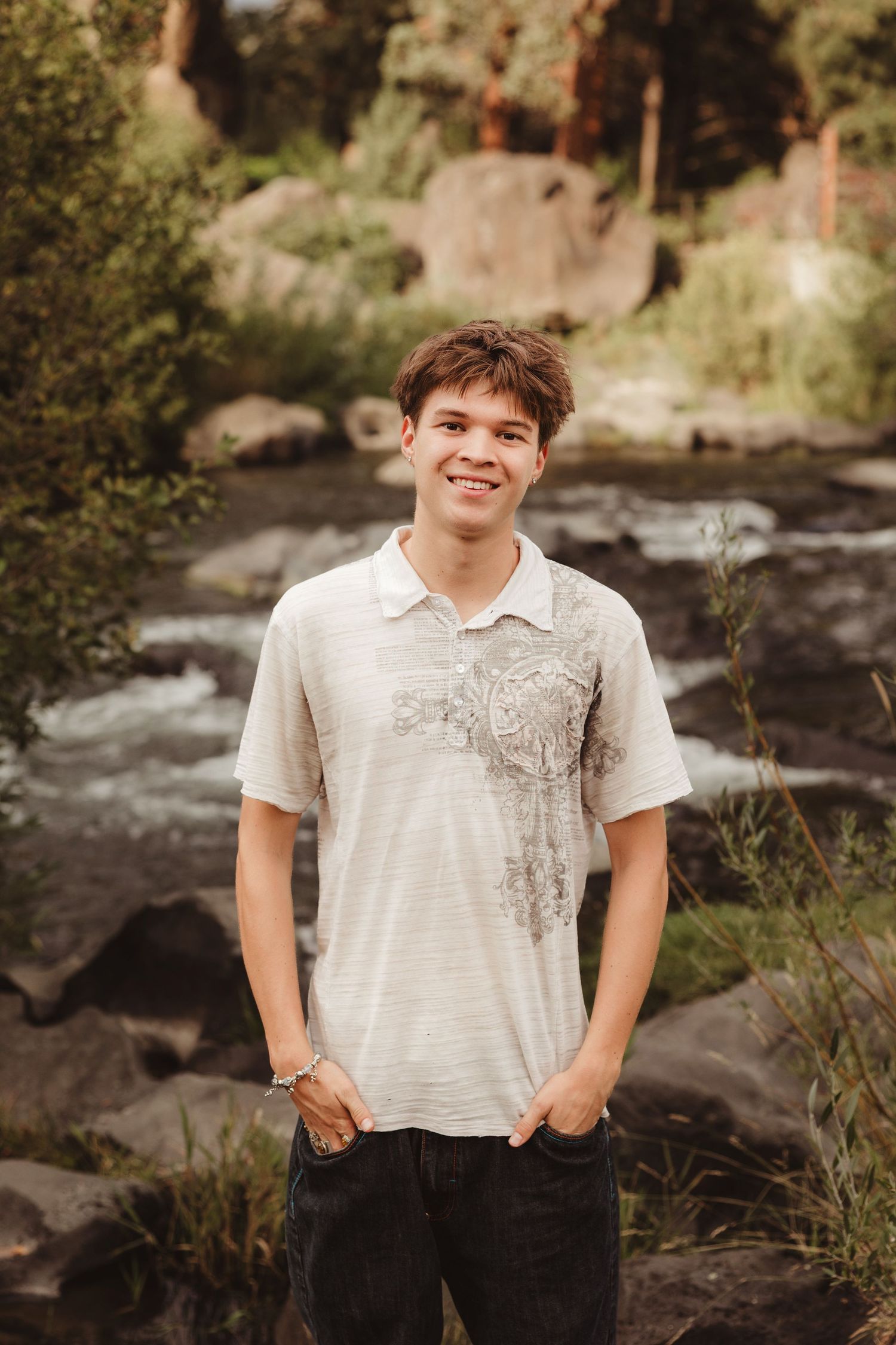 A person poses in a white shirt against a rocky river backdrop in a natural outdoor setting.