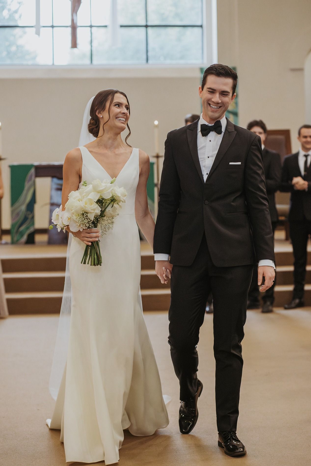 A bride and groom walk together in a church wearing a white wedding gown and black tuxedo.