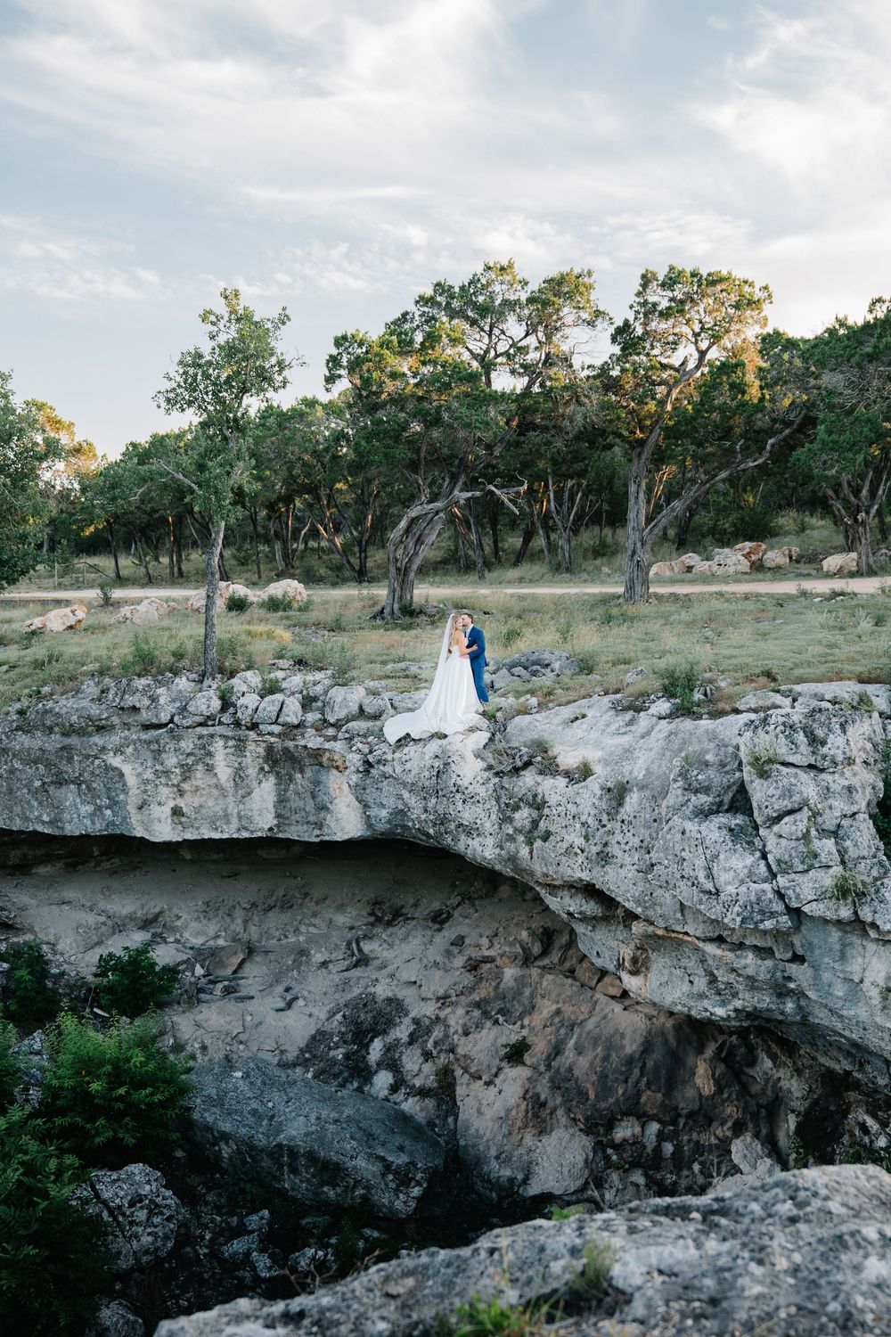 Bride and groom embracing on rocky limestone cliff edge surrounded by Texas Hill Country landscape.