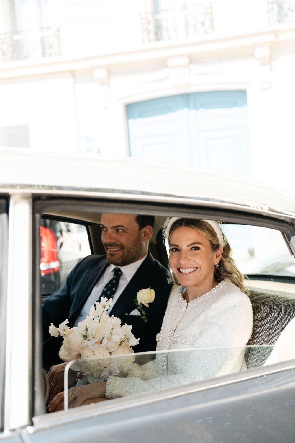 Newlywed couple smiles while sitting in vintage car after wedding ceremony, with white floral bouquet. Paris film photographer wedding.