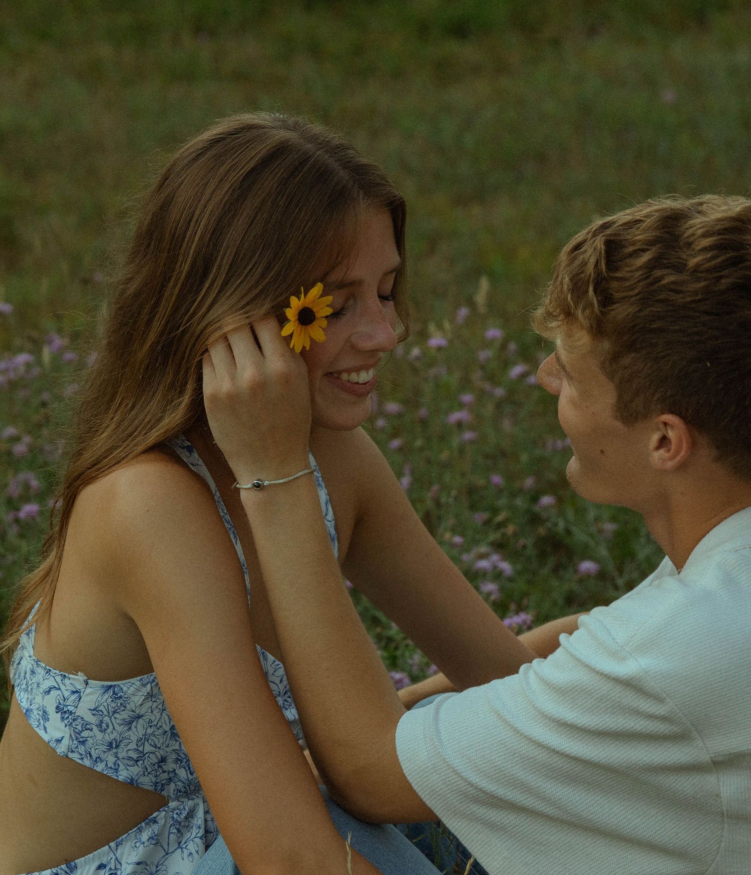 Young couple sitting in flower field, boy placing sunflower behind smiling girls ear.
