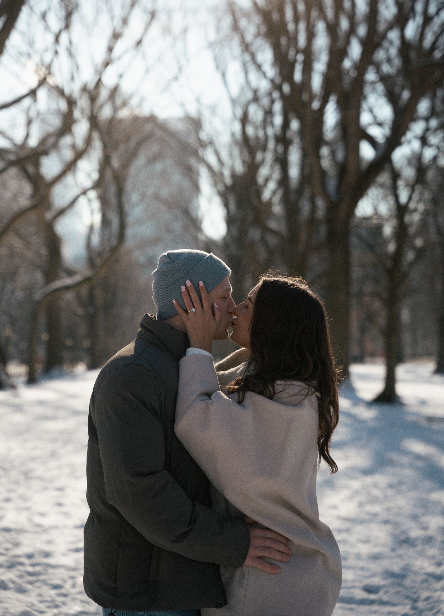 An intimate embrace captured in a snowy winter park setting with bare trees in the background.