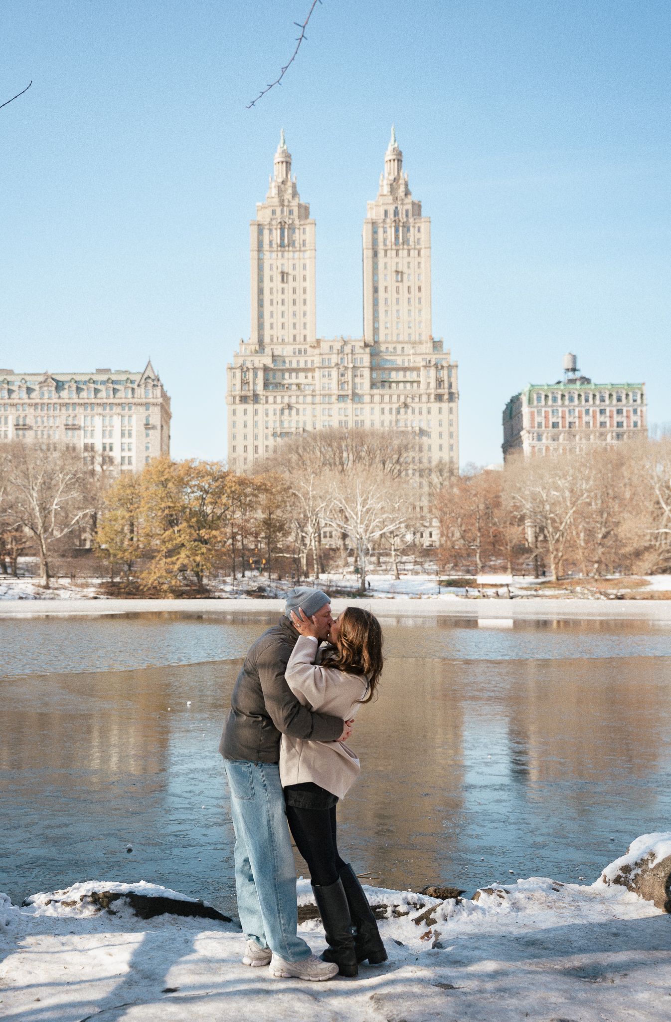 Couple shares romantic moment in snowy Central Park with iconic Manhattan buildings in background.