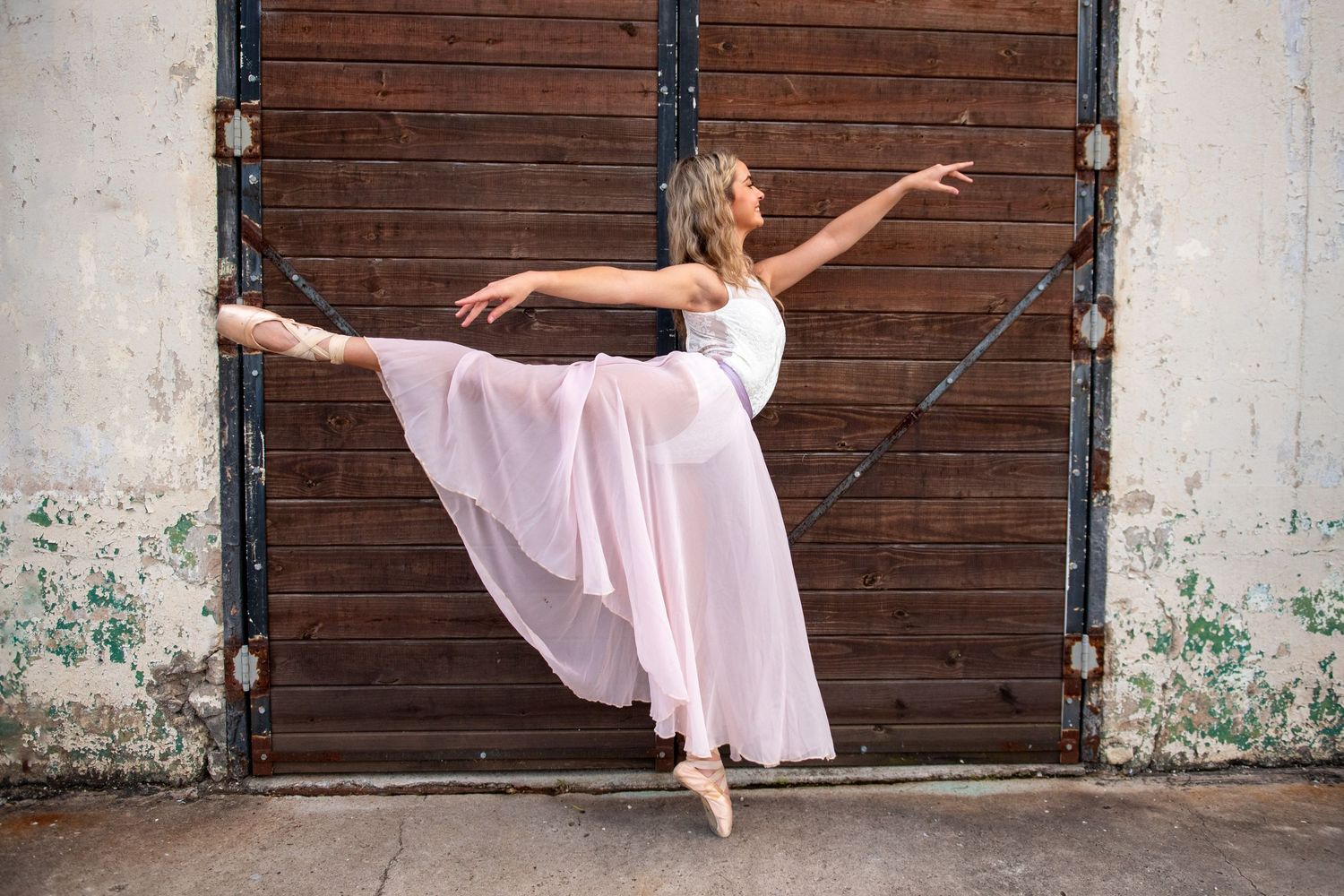 Dancer in flowing pink dress performs graceful arabesque pose against rustic wooden door and weathered wall.
