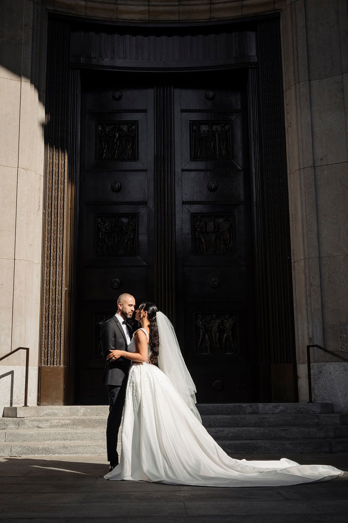 A couple in wedding attire shares an intimate moment in front of grand stone columns with dramatic lighting and shadows. Montreal, Canada