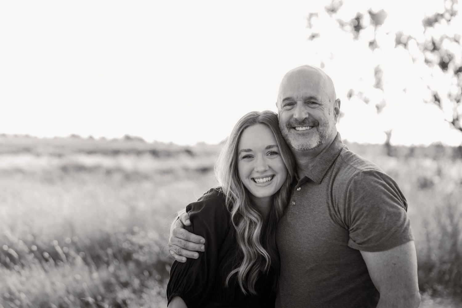Black and white portrait of two people embracing and smiling outdoors in a natural field setting.