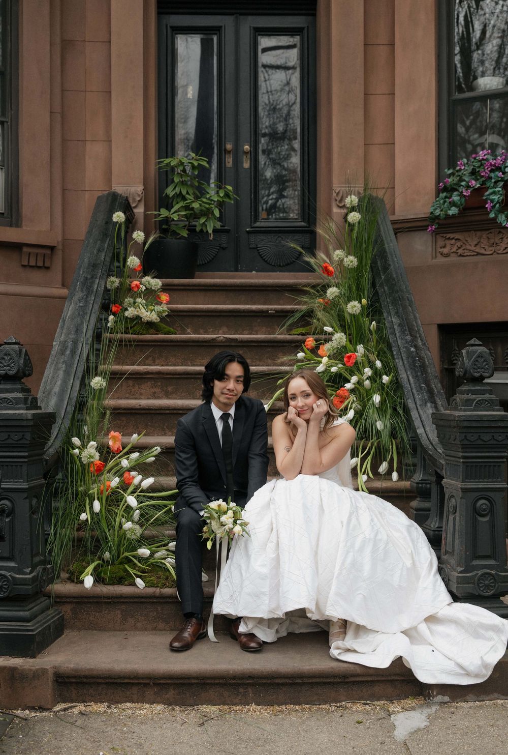 Wedding couple sits together on brownstone steps decorated with colorful flowers in an urban setting.