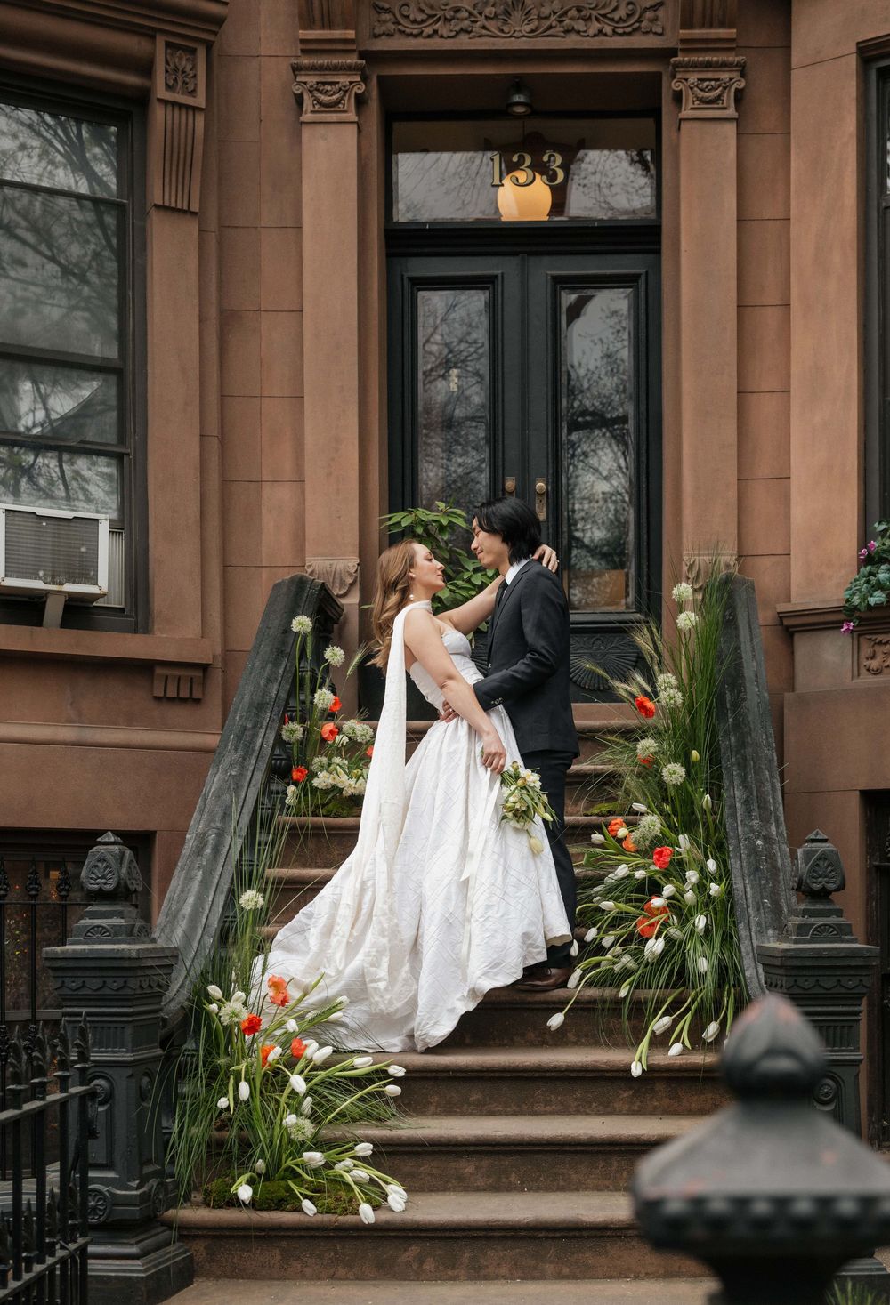 A romantic wedding couple shares tender moments on the steps of a brownstone building decorated with floral arrangements.