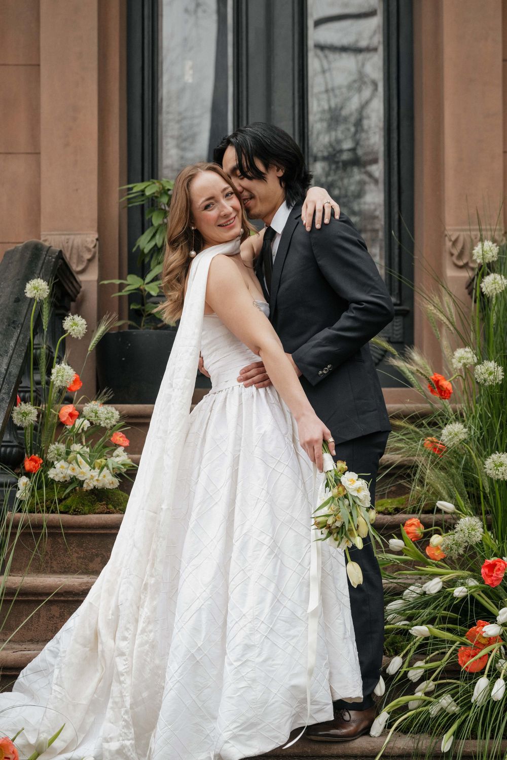 A bride in a white gown and groom in black suit embrace near vibrant red tulips and greenery outside a building.