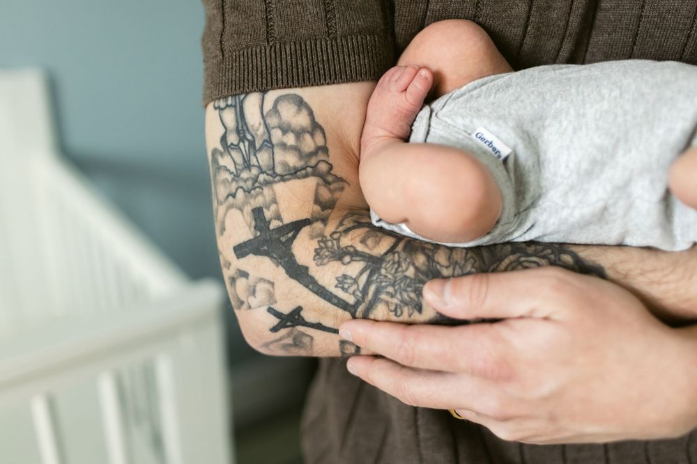 A father wearing a brown short sleeve sweater with a cross tattoo cradles his newborn baby wearing a soft gray onesie in the baby's nursery