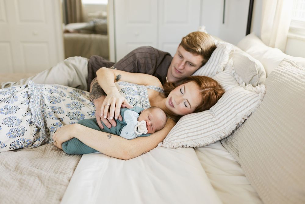 Parents lay down on a cream colored bed with their newborn son snuggled beside them for newborn photos. Concord MA NH newborn photographer.