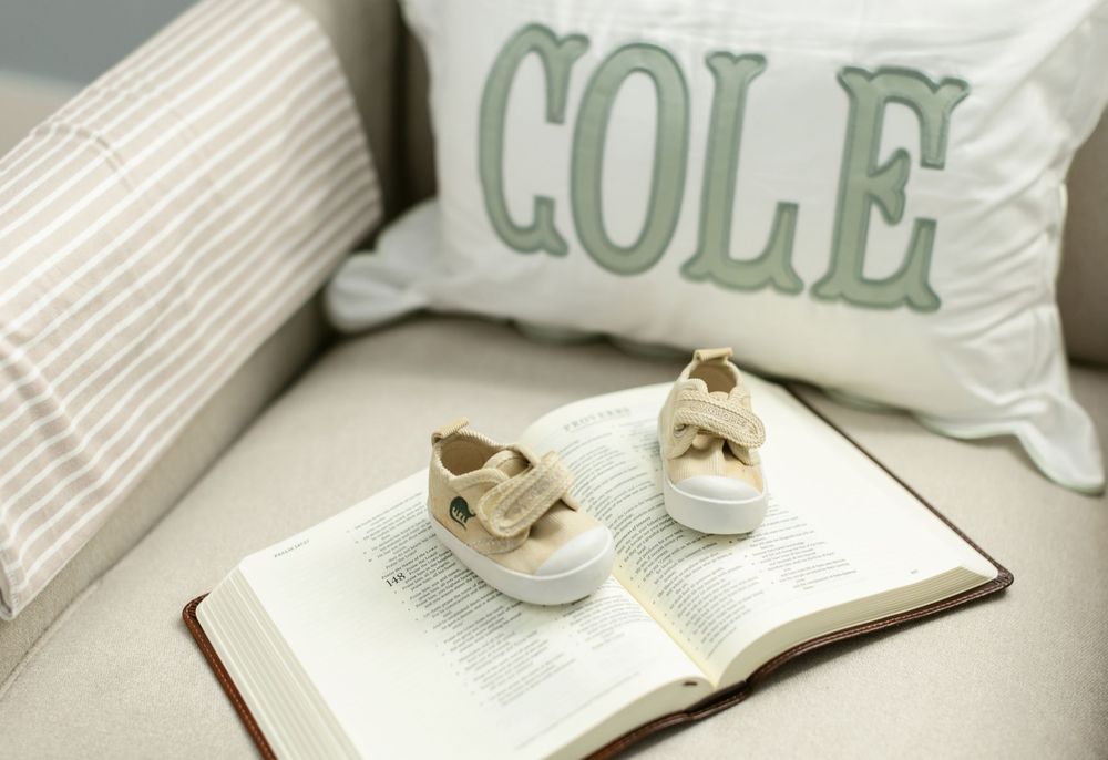 A baby pillow with the name Cole on it lays in a crib with cream newborn baby shoes on an open Bible. Portsmouth newborn photographer