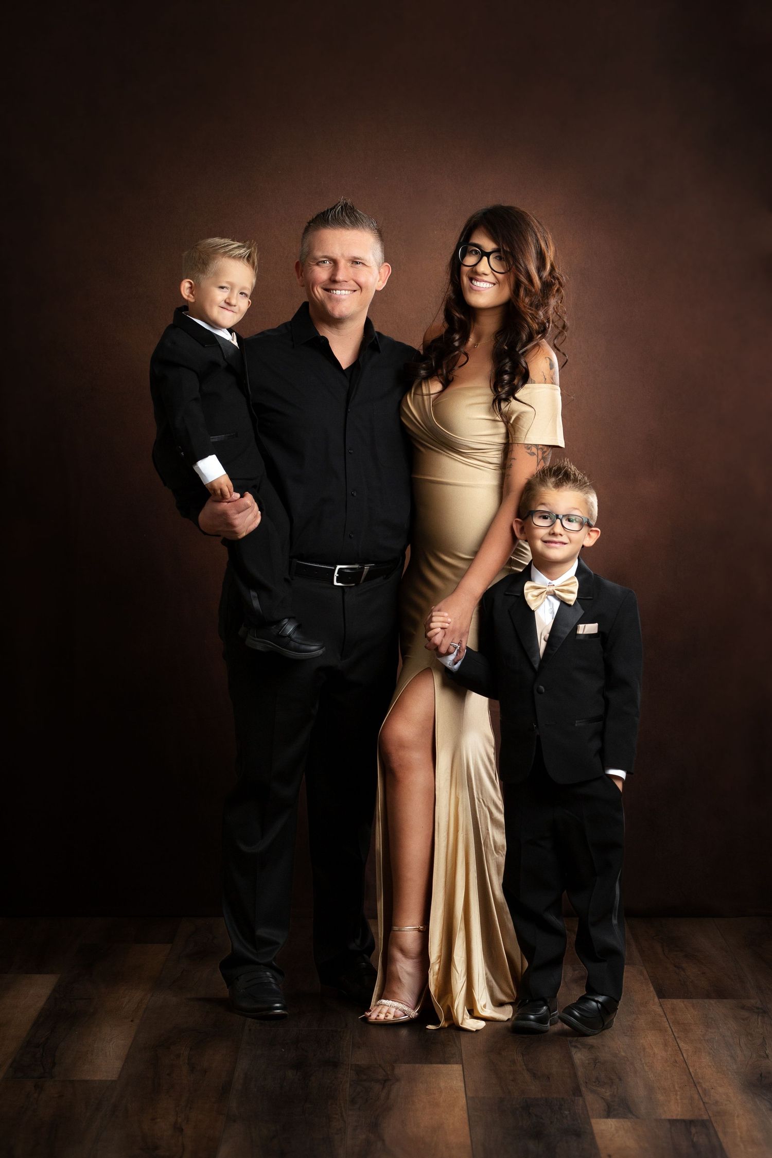 Elegant family portrait in formal attire standing together against dark brown backdrop on hardwood floor.