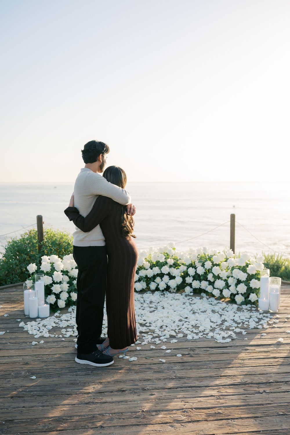 A romantic couple sits on a wooden deck overlooking the ocean with white floral arrangements at sunset.