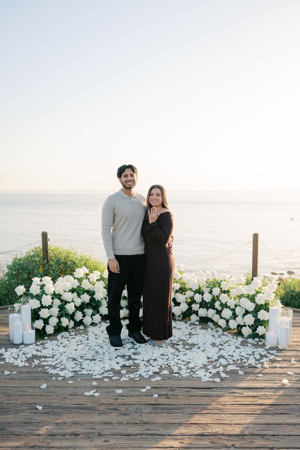 A couple stands on a wooden deck by the ocean surrounded by white floral arrangements and scattered petals at sunset.