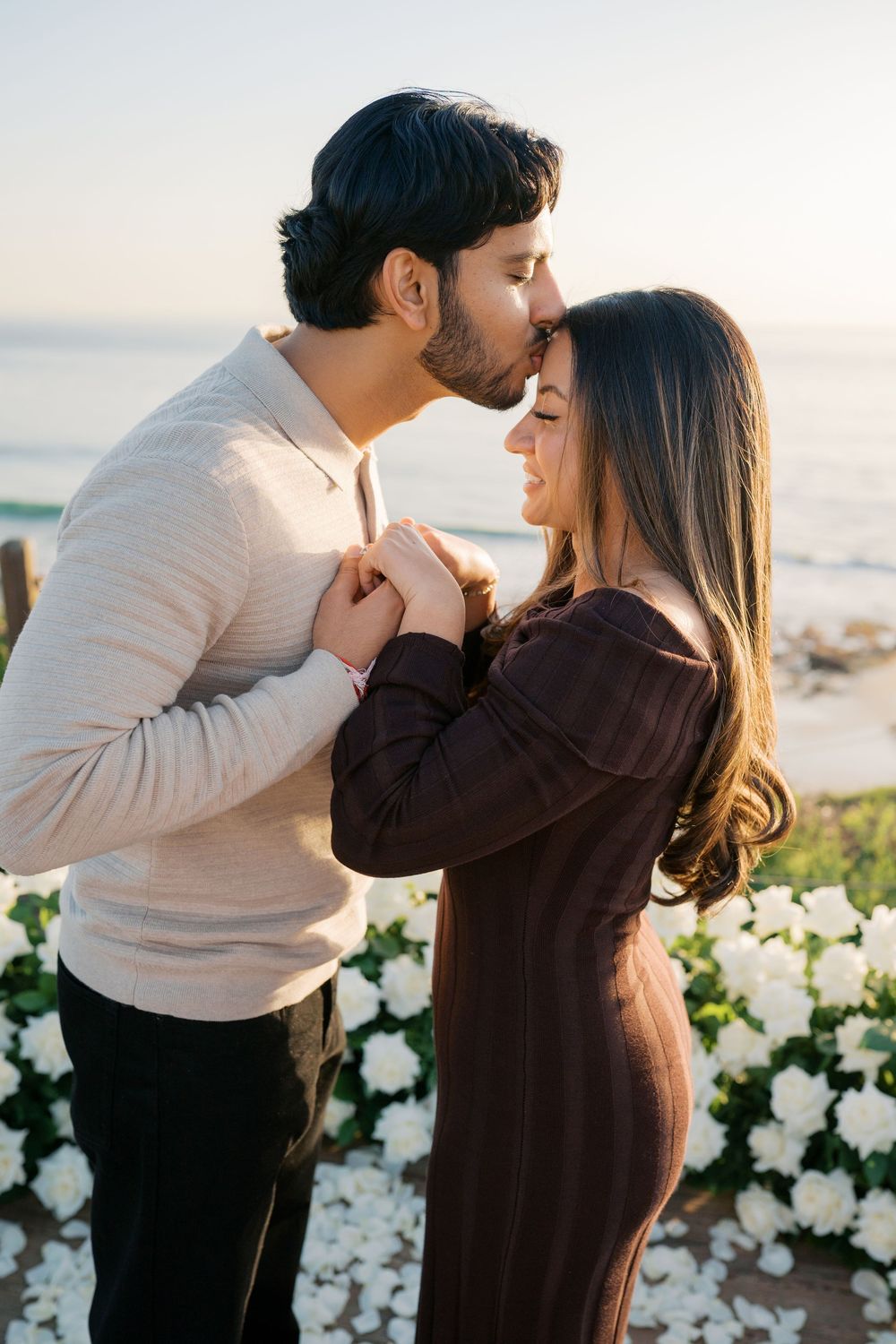 A romantic couple shares an intimate moment at sunset by the ocean with white flowers in the foreground.