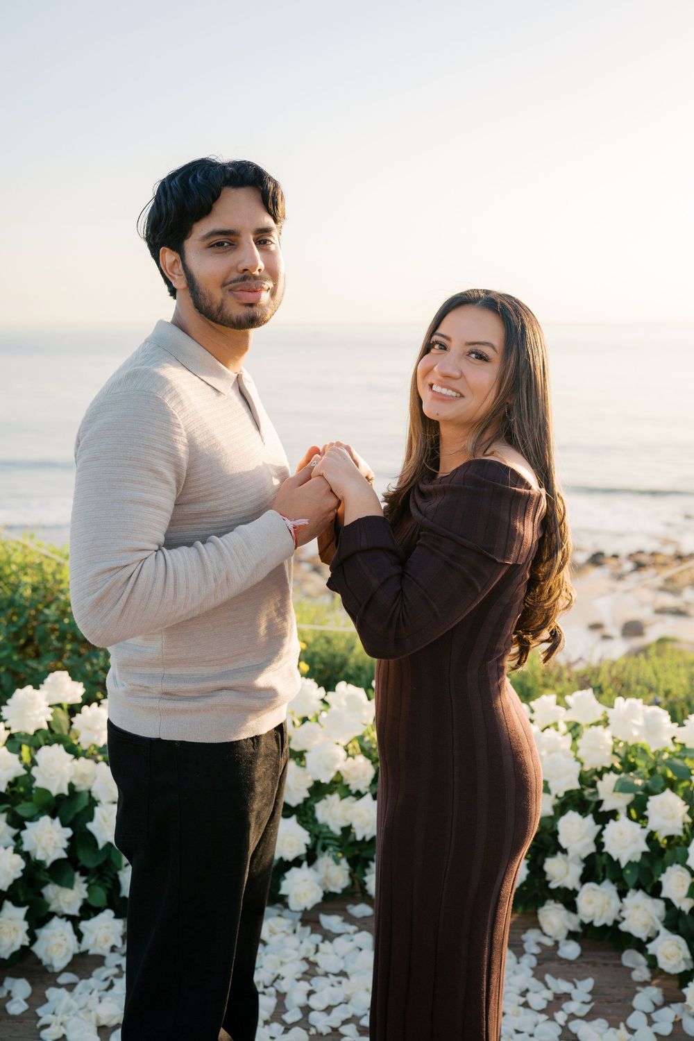 A couple holds hands and smiles while standing by white flowers near the ocean at sunset.