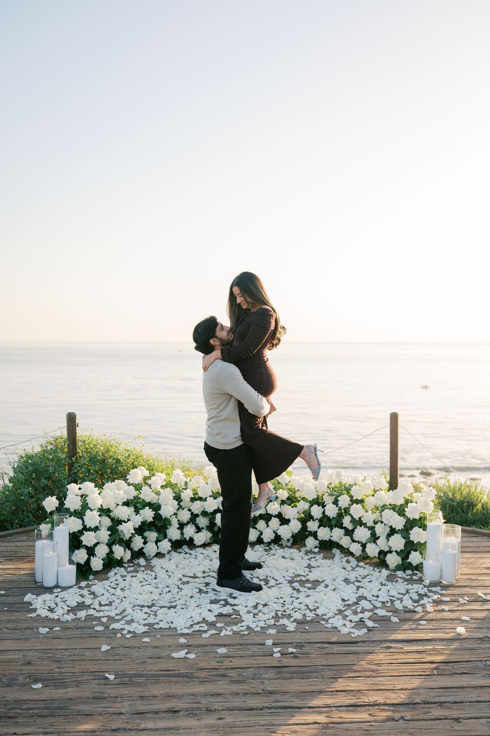 A romantic proposal moment on an oceanside deck decorated with white flowers and scattered rose petals.