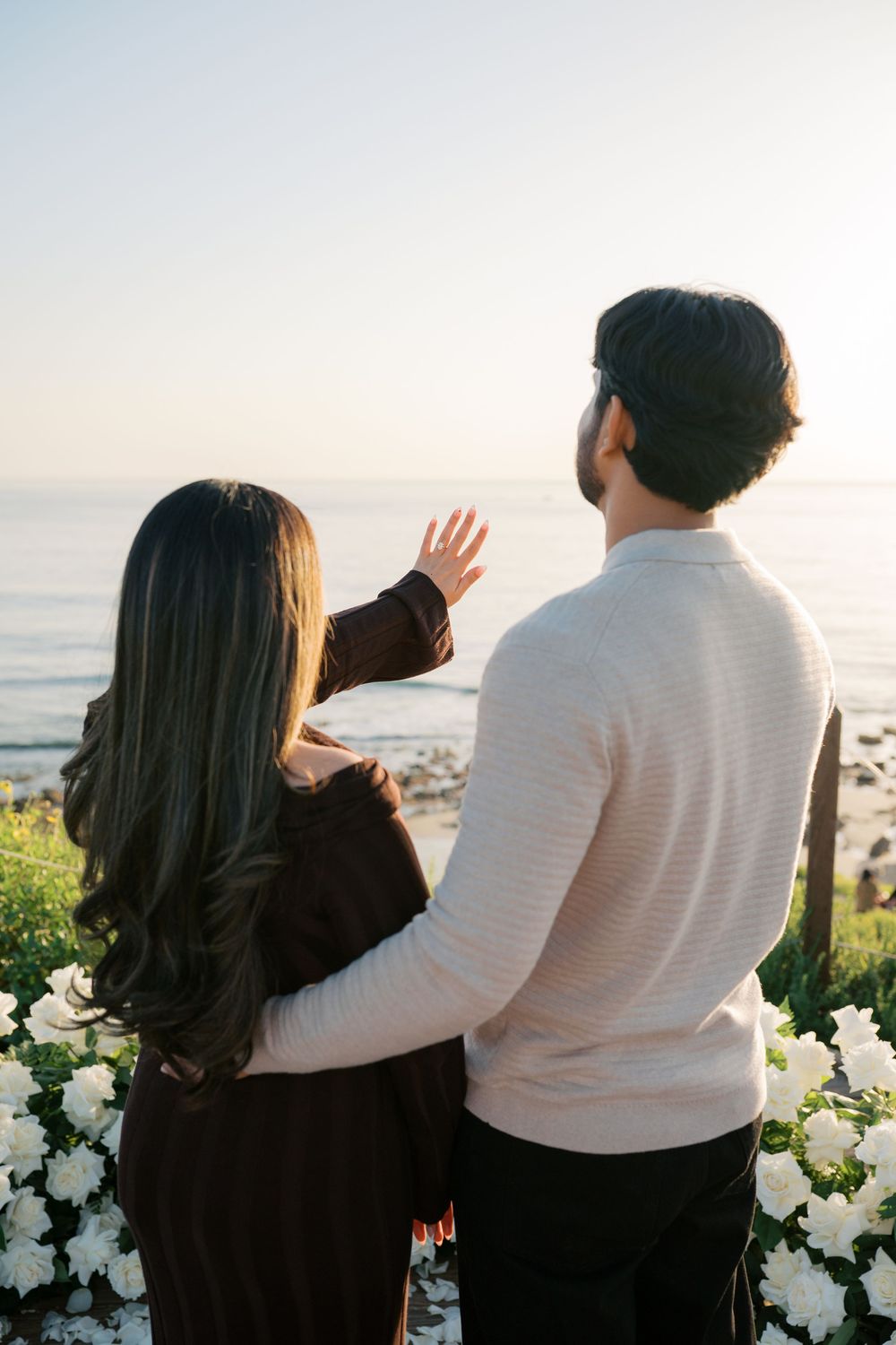 Couple stands together overlooking ocean view at sunset with white flowers in foreground.