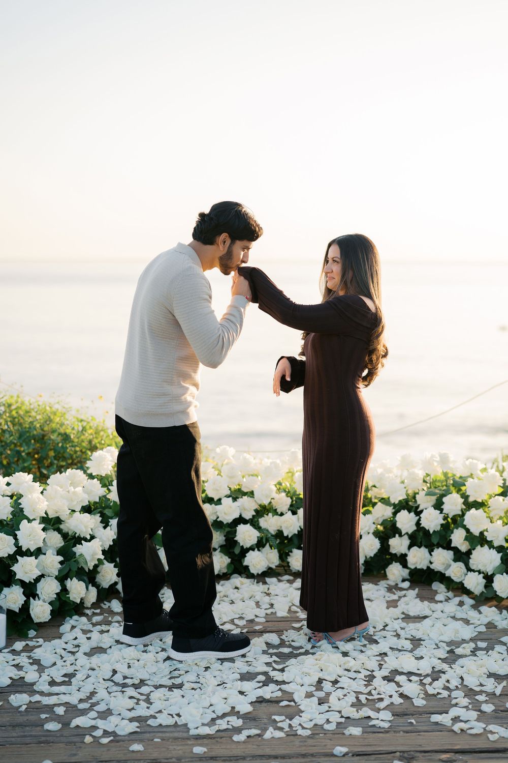 A couple dances together at sunset surrounded by white rose petals and floral arrangements on an outdoor terrace.