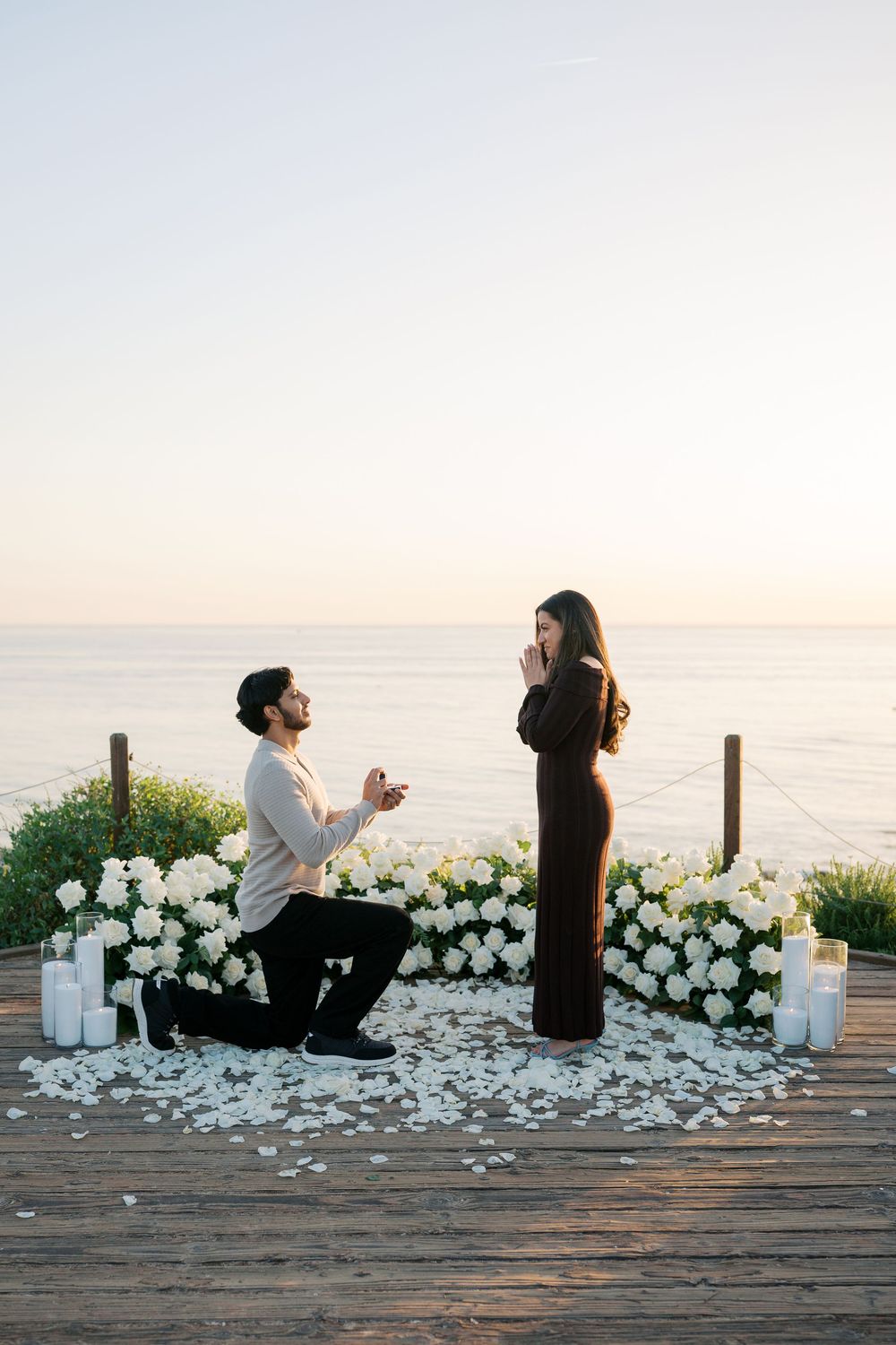 A romantic beach proposal scene with white rose petals scattered on a wooden boardwalk at sunset.