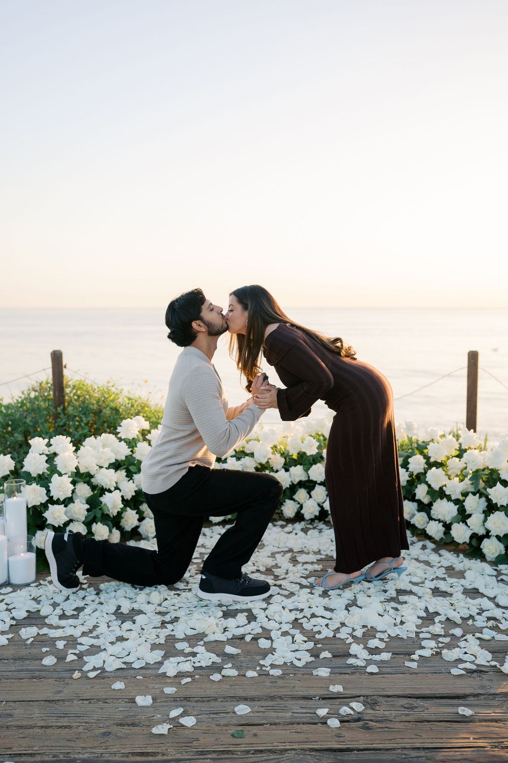 Couple shares intimate moment on wooden deck surrounded by scattered white rose petals at sunset.