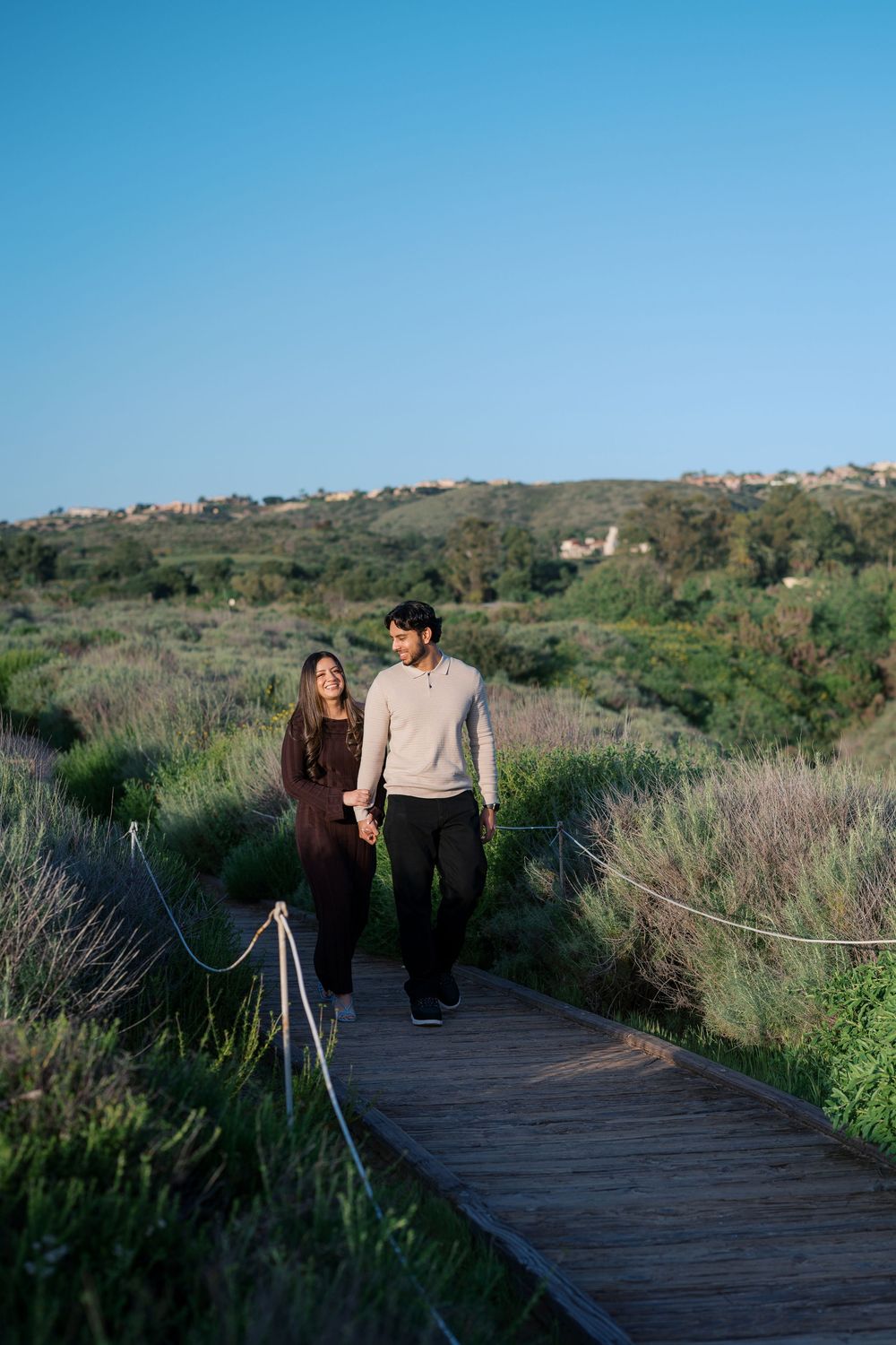 Two people walking along a wooden boardwalk path through coastal wetlands on a sunny day.