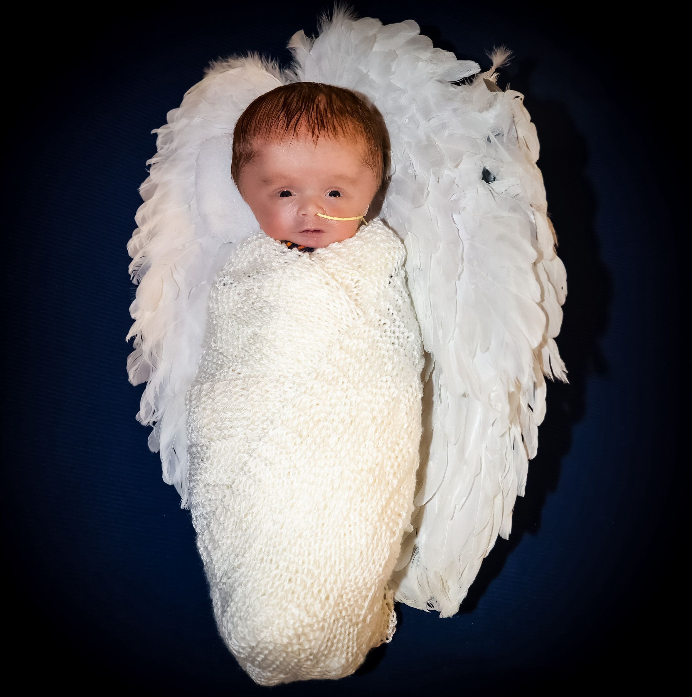 A sleeping newborn wrapped in white swaddle with angel wings against dark background.