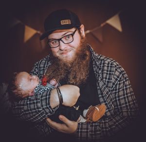 Dark moody family photoshoot with baby and parents in casual attire against black background with banner decorations.