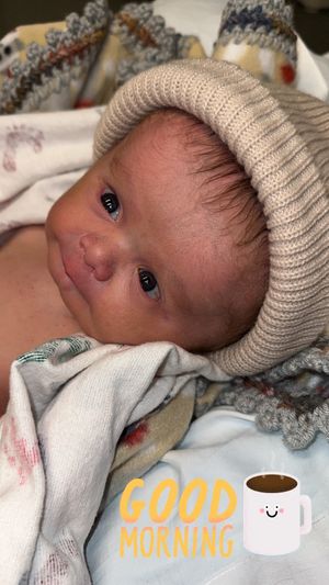 A baby wearing a knit beanie lies on patterned bedding with a "Good Morning" text overlay and coffee cup emoji.