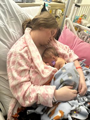 Caregiver in pink patterned robe tenderly holds sleeping newborn in hospital setting.