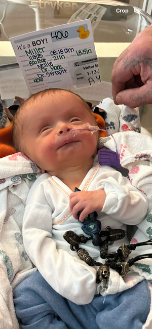 Newborn baby in hospital wearing white and blue outfit with medical tags visible.