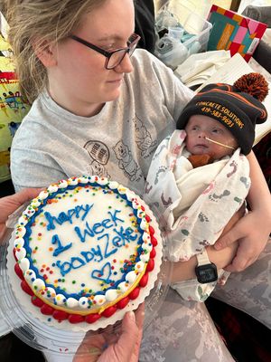 A celebratory cake reading 'happy 1 week baby' next to bundled newborn in winter hat.