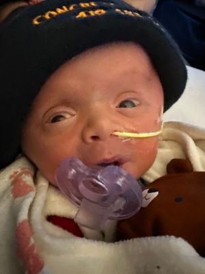 Baby with pacifier wearing dark hat rests against plush toy.