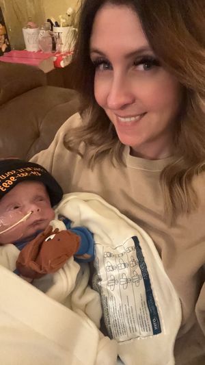 Mother in cream colored sweater cuddles with newborn while holding medical supplies.