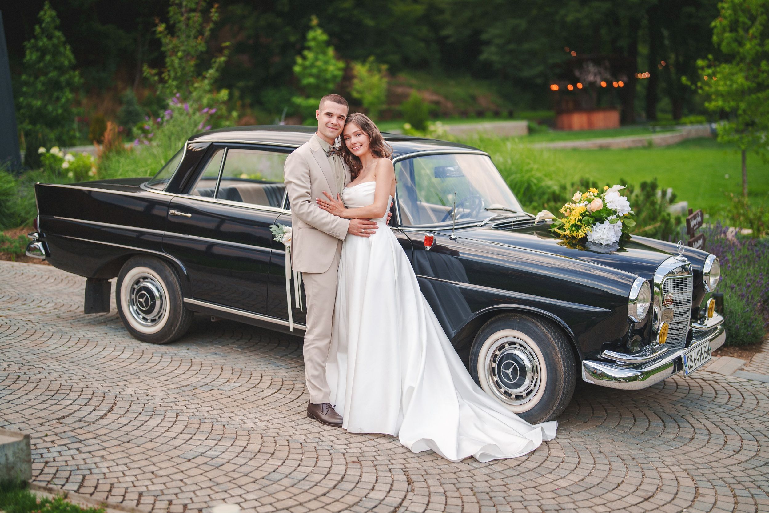 A vintage black luxury car serves as an elegant backdrop for wedding photos on a cobblestone driveway.