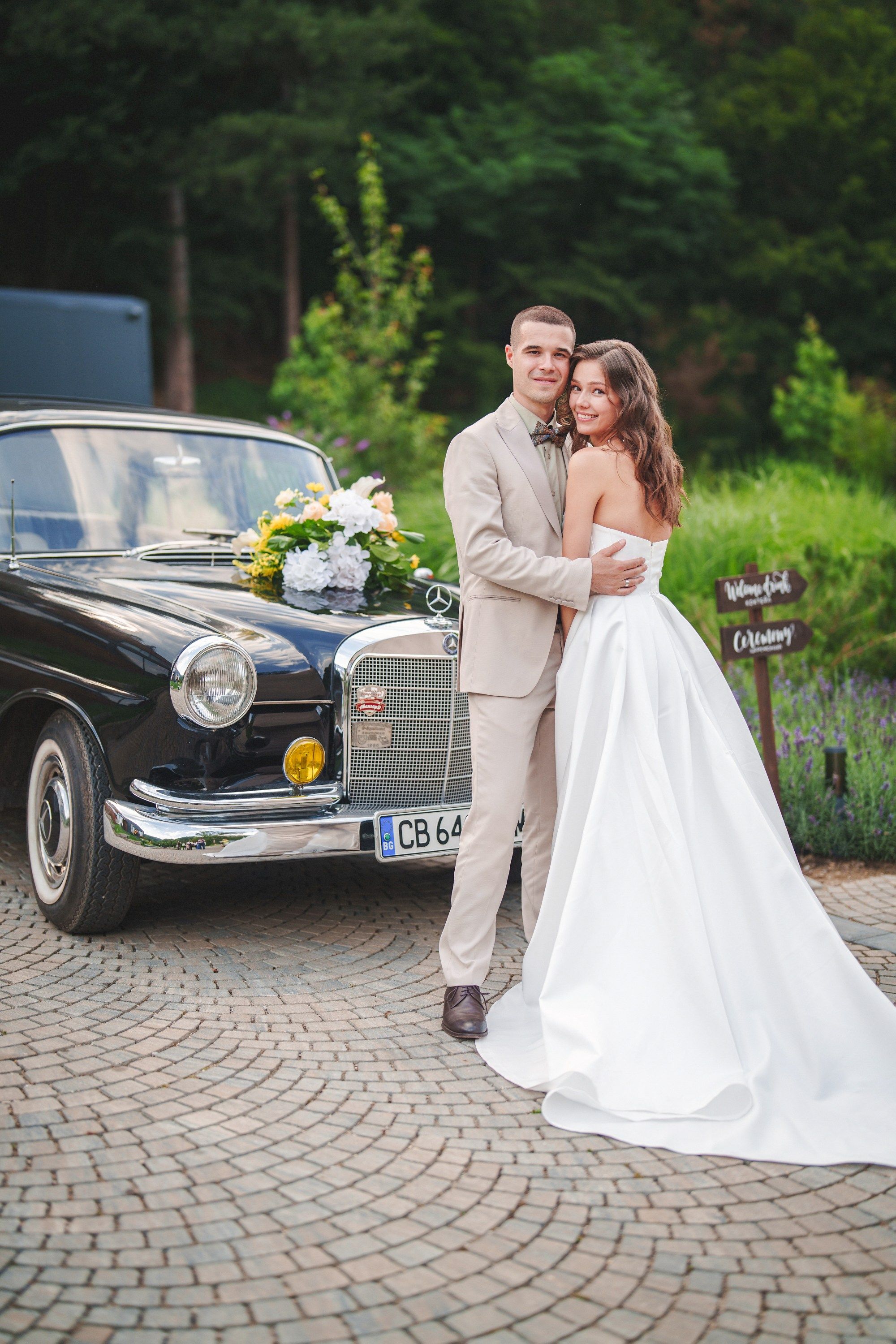 A couple poses with a classic vintage car on a cobblestone driveway during their elegant outdoor wedding celebration.