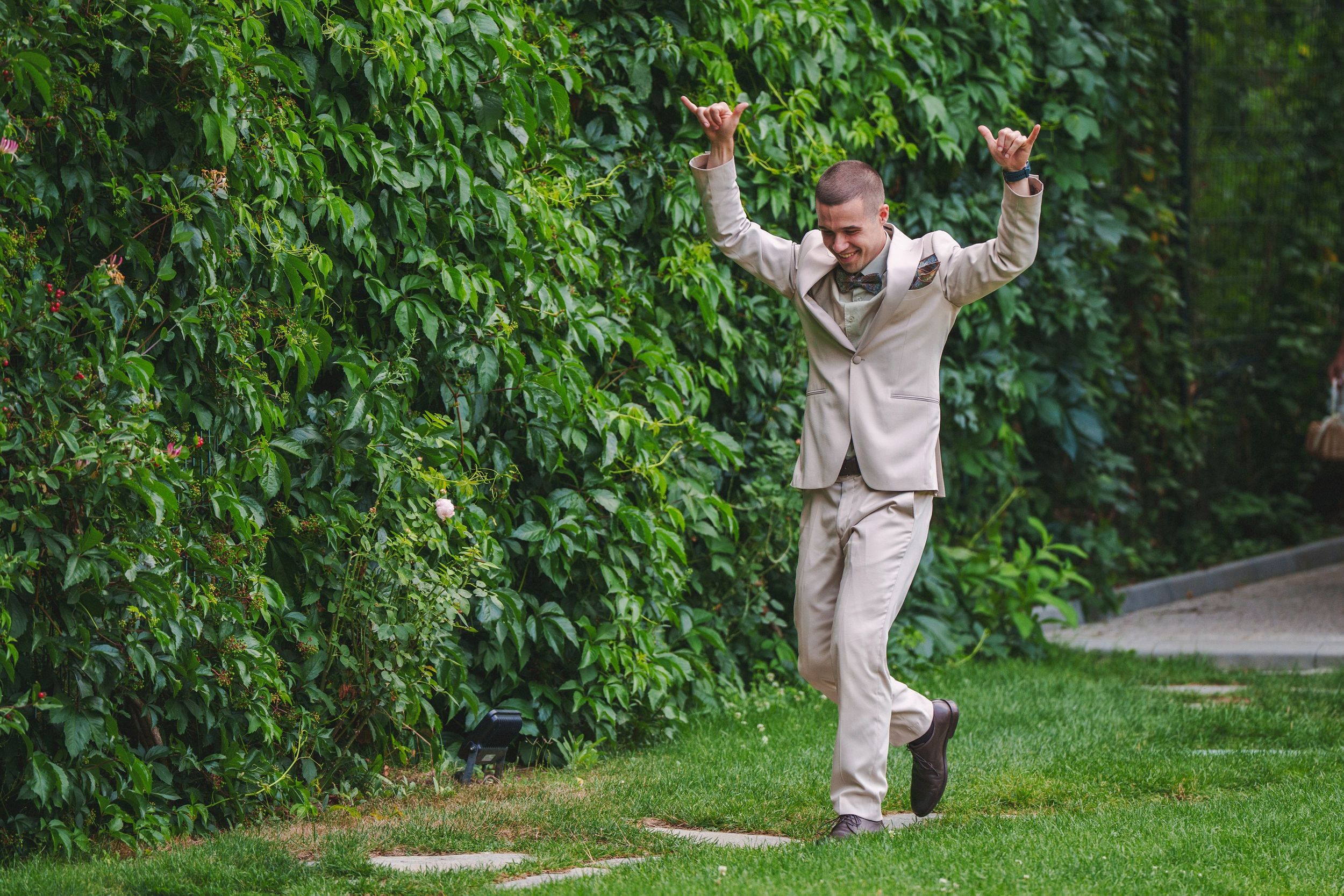 A group in beige suits poses playfully on stepping stones in a lush garden setting.