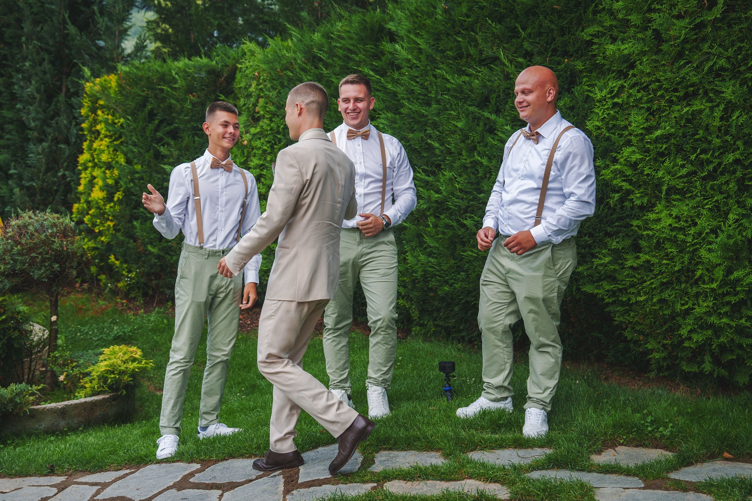Wedding party members in light colored suits interact casually on stone path surrounded by greenery.