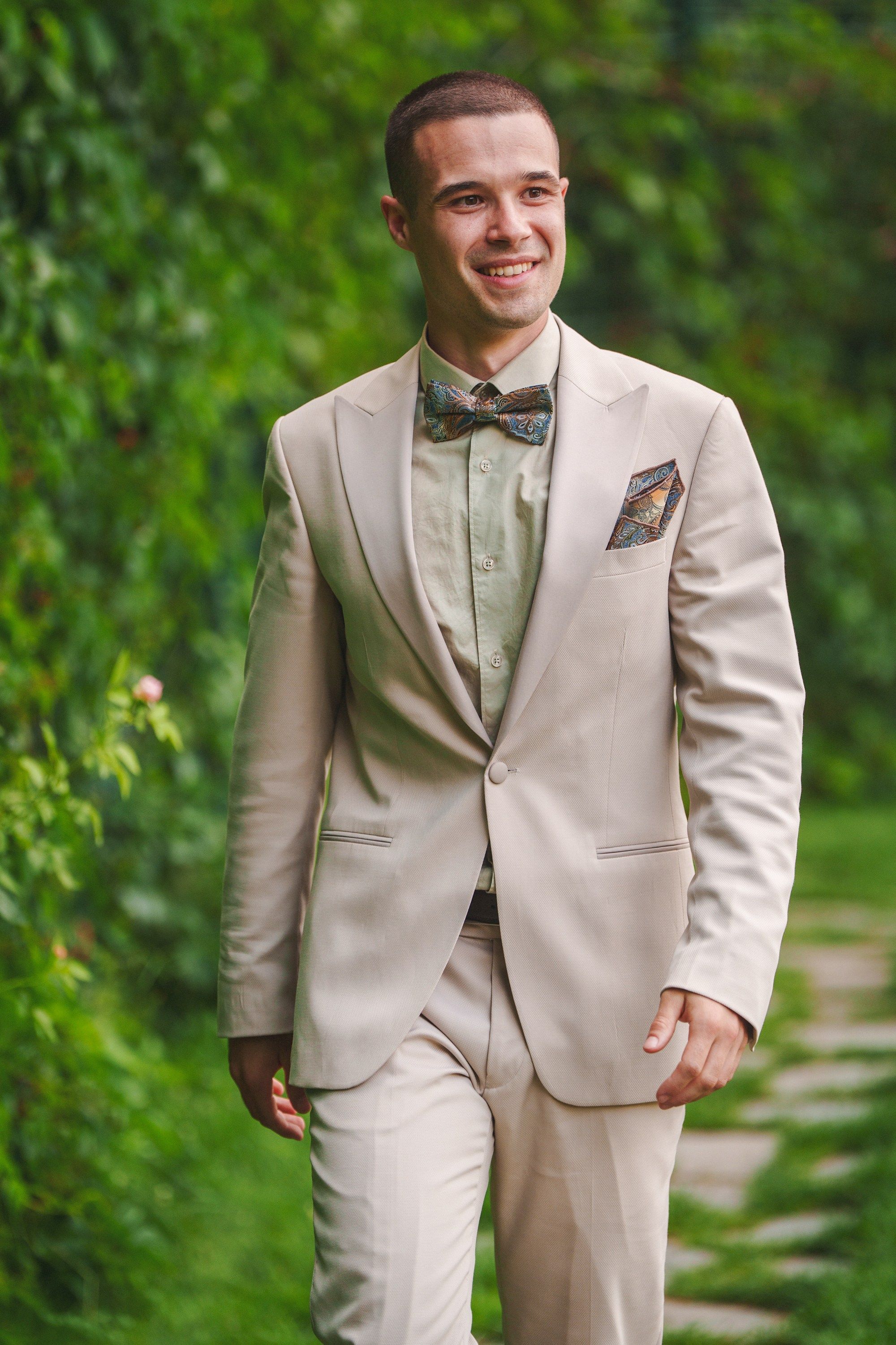 A person in a light beige suit and bowtie poses outdoors against a green natural background.