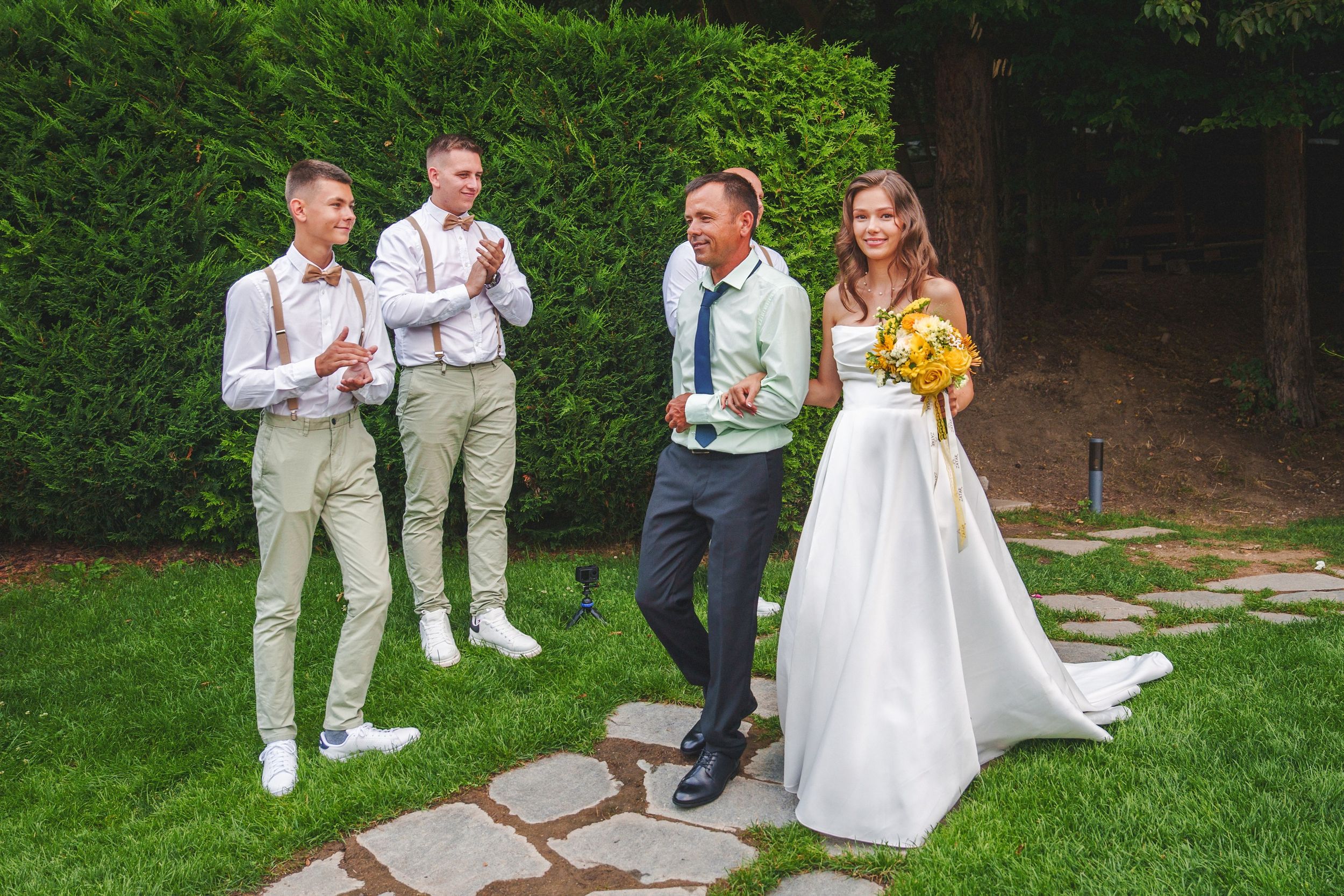 Wedding party members in light colored attire gather for outdoor photos in a lush garden setting.