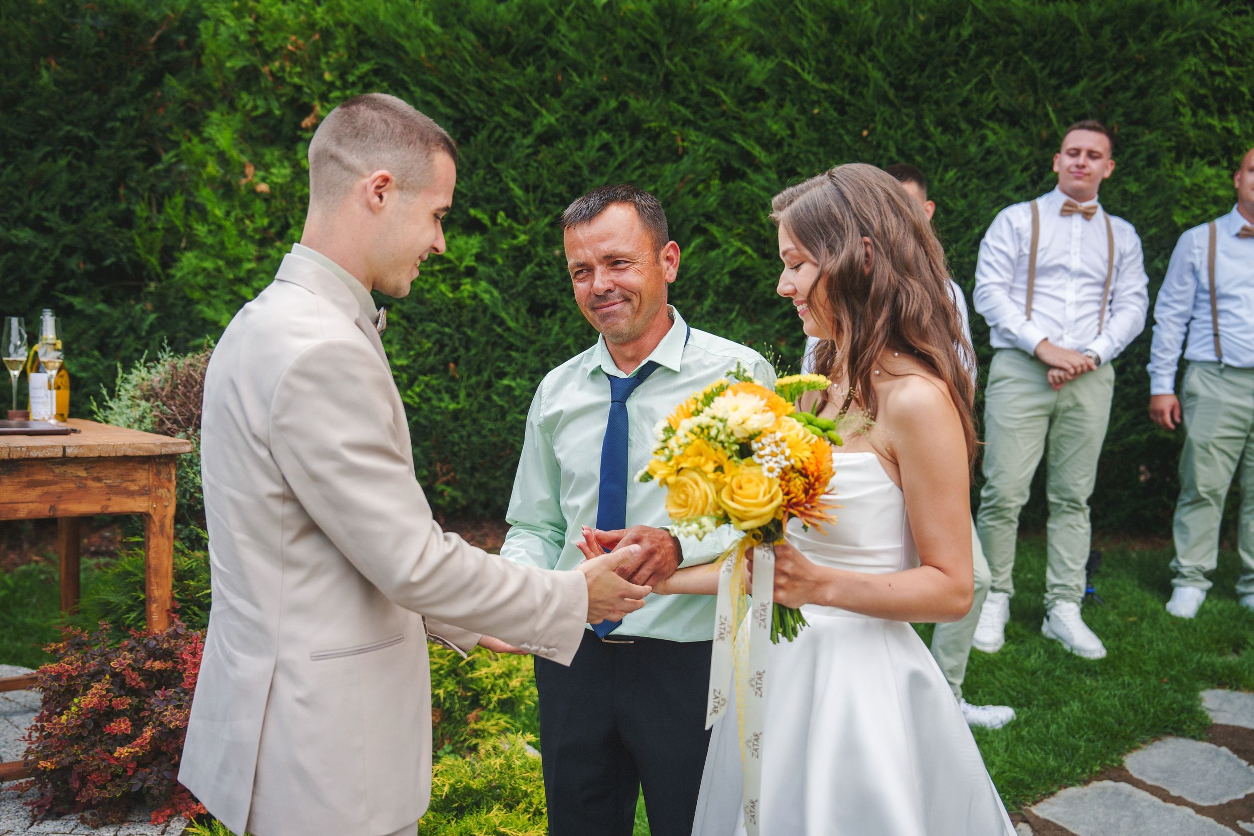 Wedding party in formal attire poses for candid photos in a lush garden setting during a summer celebration.