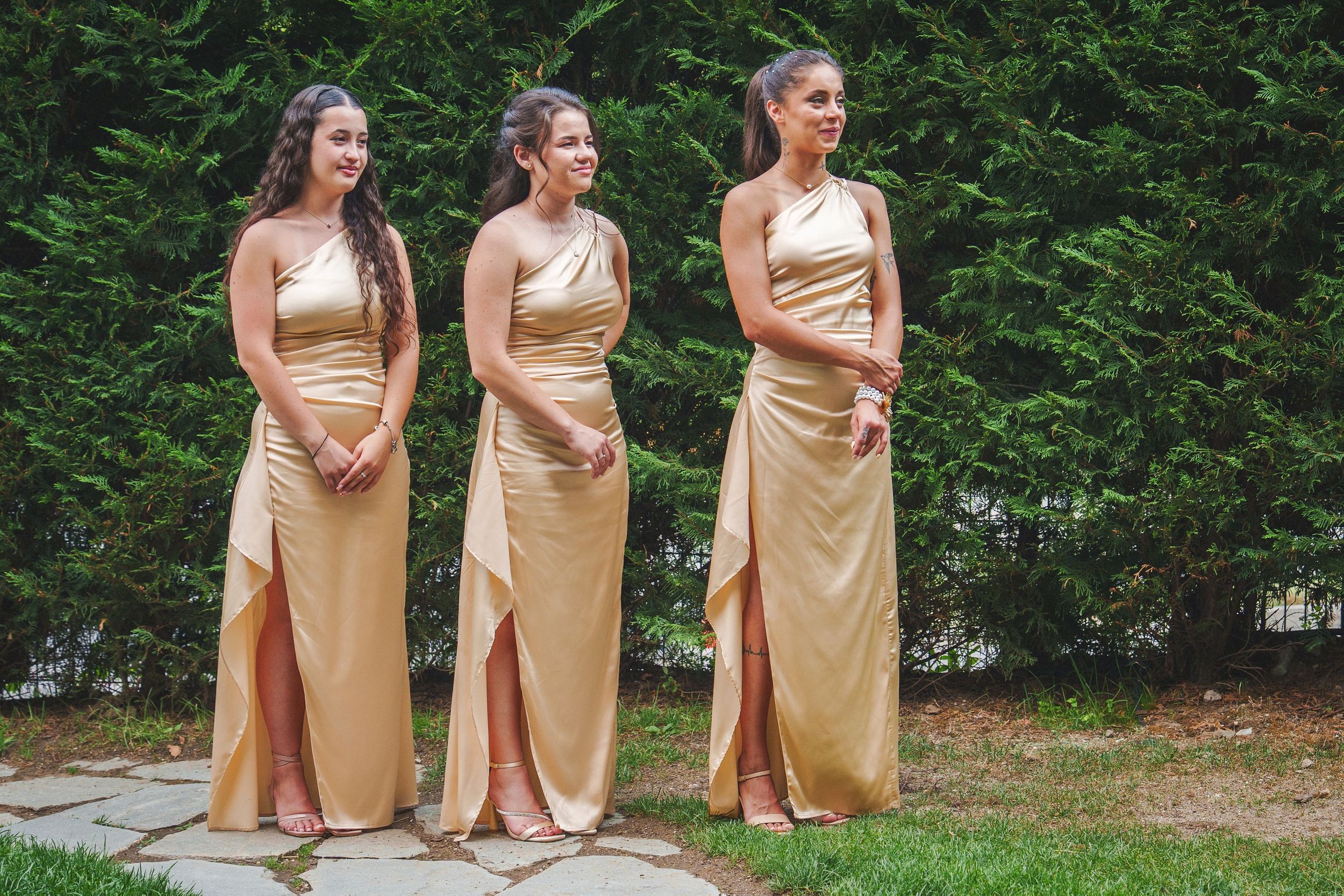 Three bridesmaids in champagne-colored dresses pose outdoors with matching bouquets in a garden setting.