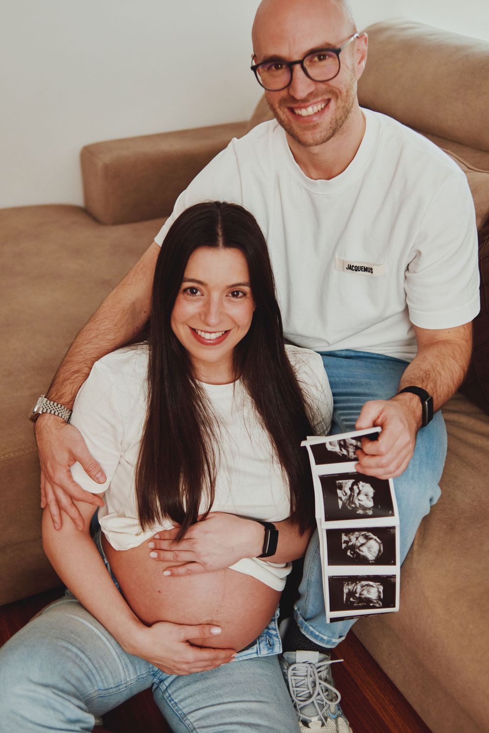 Happy couple sitting on sofa holding ultrasound scans, pregnant woman touching her bare belly.