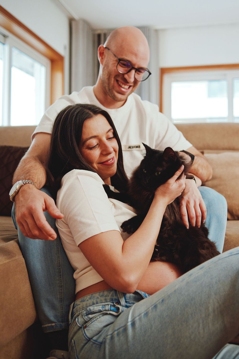 Happy couple sitting on couch petting their fluffy black cat together in a cozy living room.