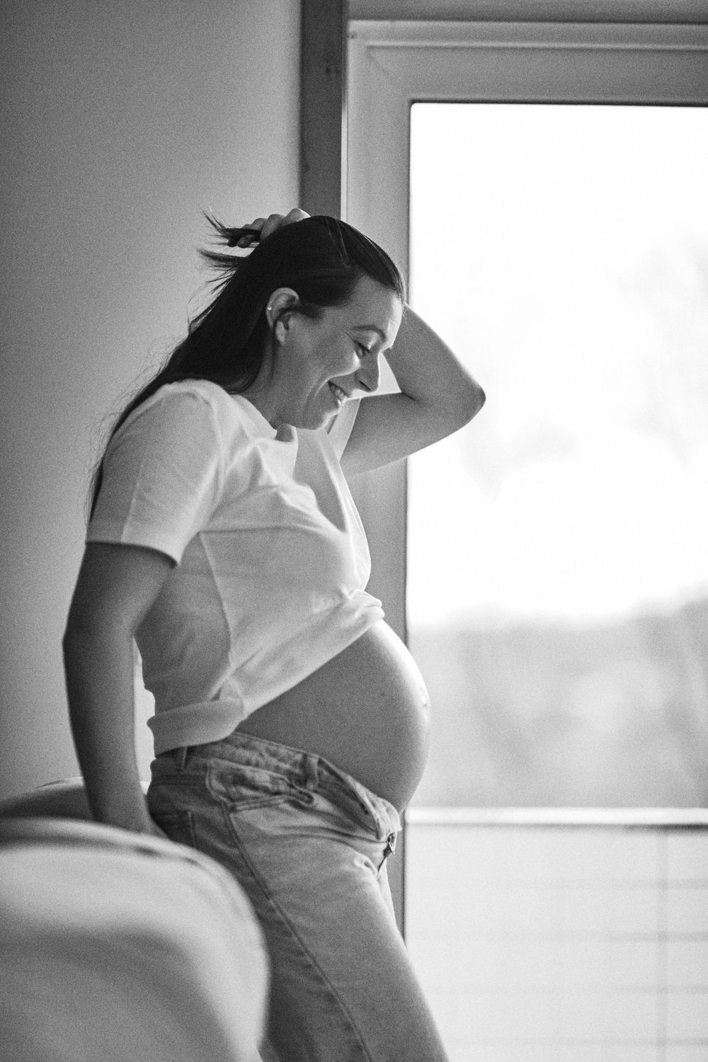 Black and white photo of smiling pregnant woman standing by window in white tee and jeans.