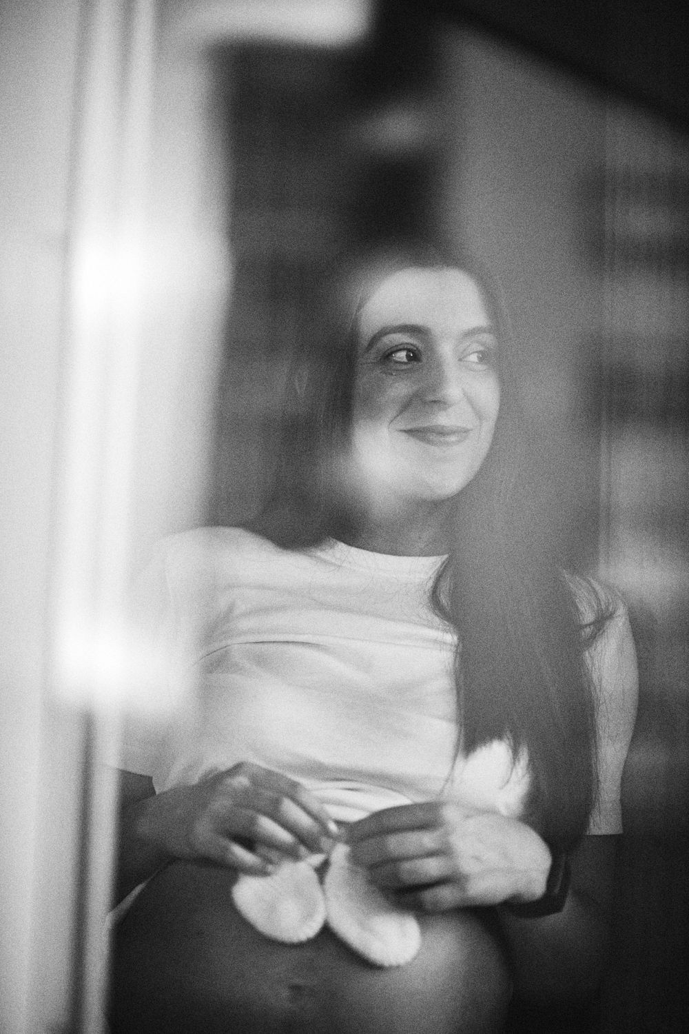 Black and white portrait of smiling pregnant woman holding baby booties, shot through glass.