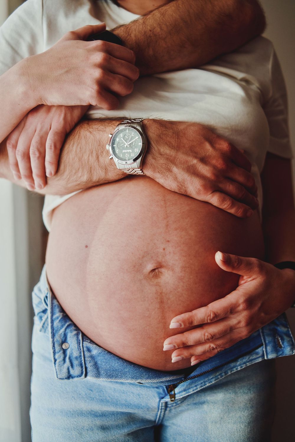 Close-up of couple embracing pregnant belly, mans hand wearing silver watch wrapped around bump.