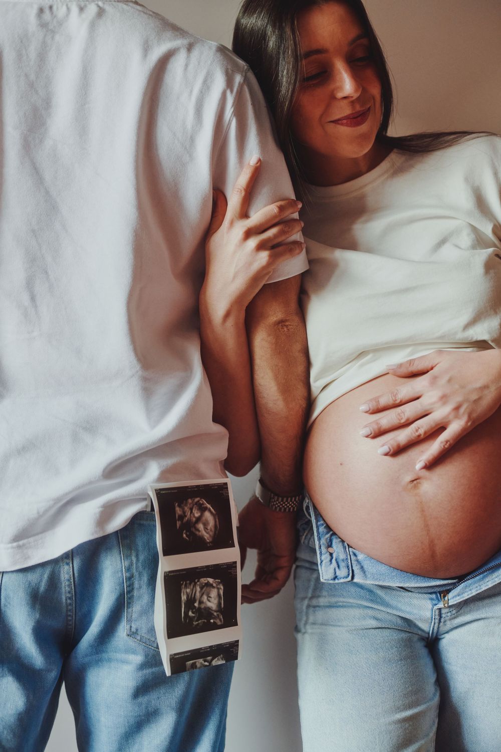 Pregnant woman leaning on partner holding ultrasound photos while she cradles her baby bump.